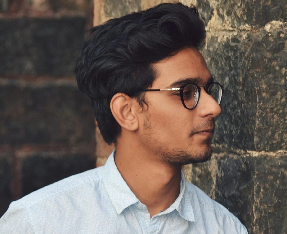 A stylish young man in glasses leans against a brick wall wearing a casual light blue shirt.